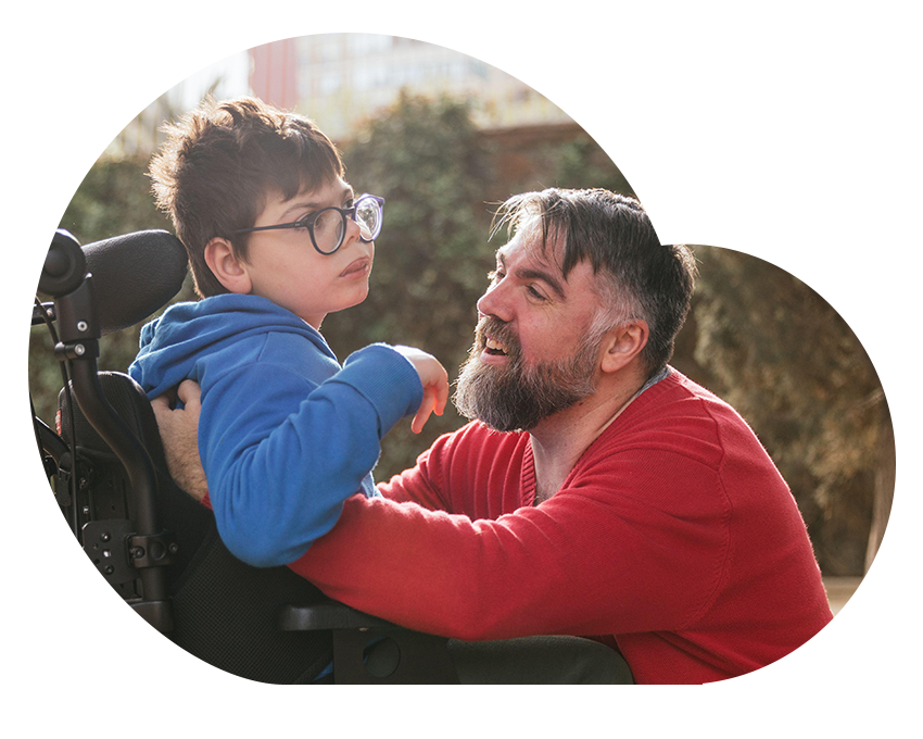 A father crouches beside his young son in a wheelchair, smiling and offering support outdoors.